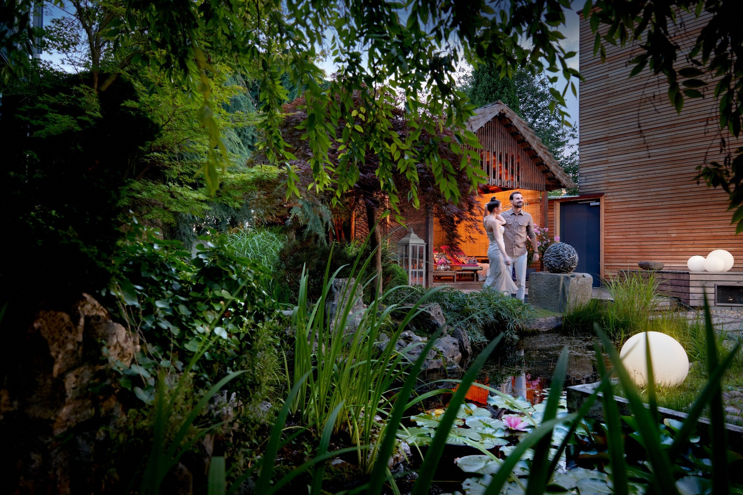 Paar spaziert durch Ganesha's Garten, umgeben von üppigem Grün, Teich mit Seerosen und einer gemütlich beleuchteten Holzhütte im Hintergrund.